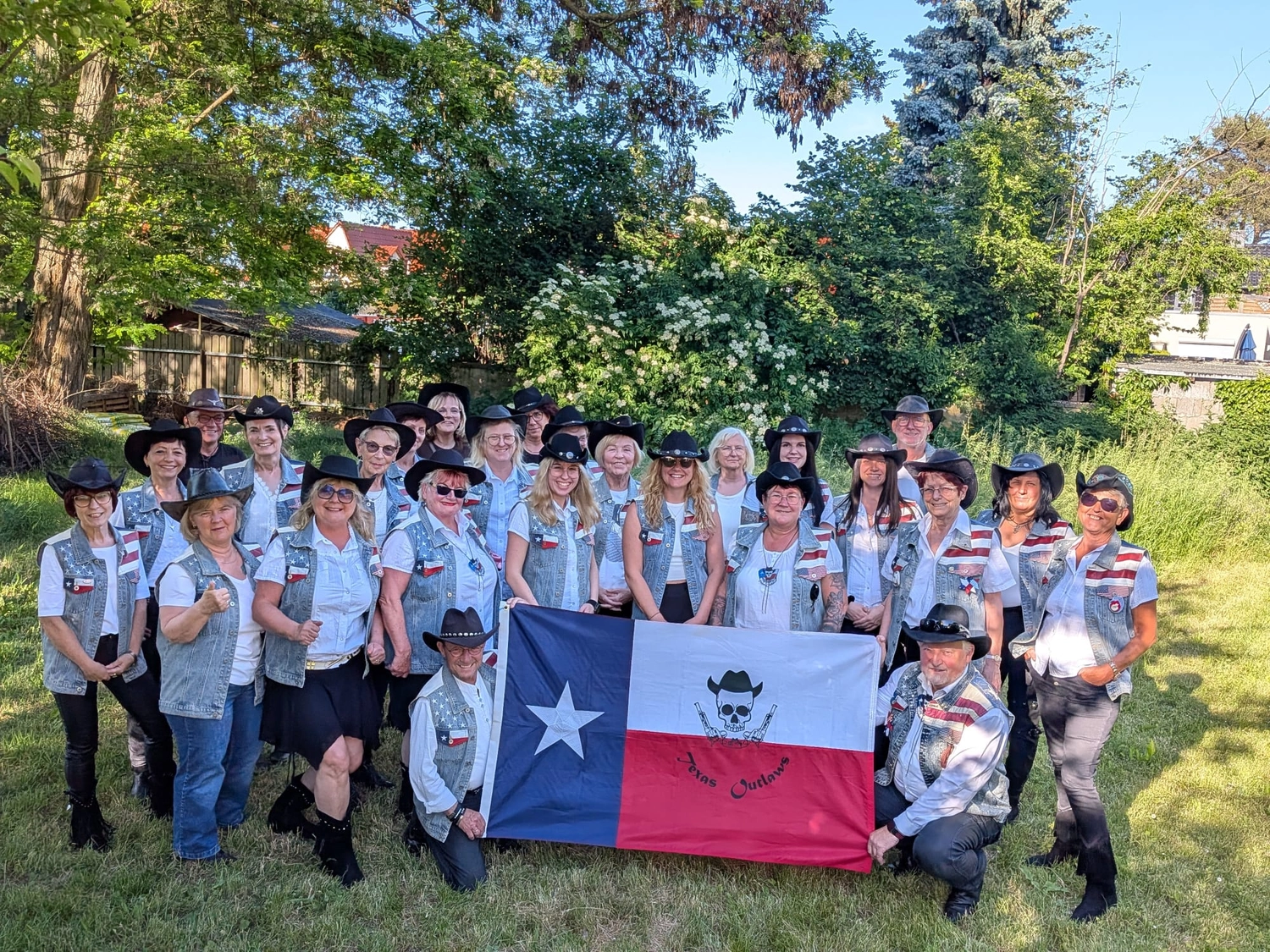 Texas Outlaws - LINE DANCE CLUB - Eine Gruppe von Line-Dancern in Cowboyhüten und passenden Jeanswesten posiert im Freien und hält eine Texas-Flagge mit einem individuellen Totenkopf-Logo.