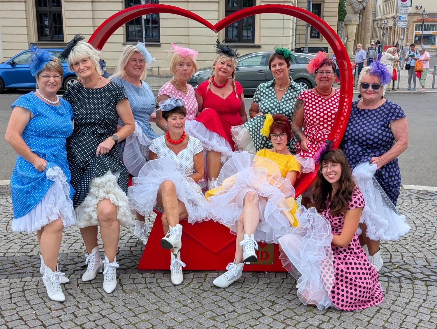 Texas Outlaws -  Line dance club - Frauen in Retro-Kleidern mit Polka-Dot-Muster und weißen Petticoats versammeln sich um eine große Skulptur in Form eines roten Herzens.